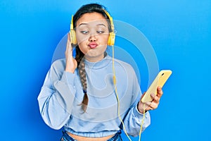 Young hispanic girl using smartphone and headphones making fish face with mouth and squinting eyes, crazy and comical