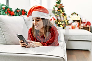 Young hispanic girl using smartphone lying on the sofa by christmas tree at home