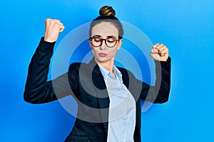 Young hispanic girl wearing business clothes and glasses showing arms muscles smiling proud