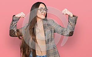 Young hispanic girl wearing business clothes and glasses showing arms muscles smiling proud