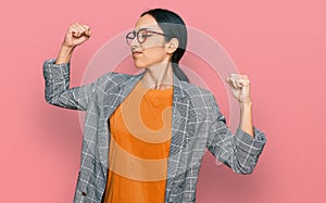 Young hispanic girl wearing business jacket and glasses showing arms muscles smiling proud