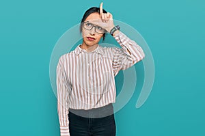 Young hispanic girl wearing casual clothes and glasses making fun of people with fingers on forehead doing loser gesture mocking