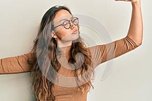 Young hispanic girl wearing casual clothes and glasses showing arms muscles smiling proud