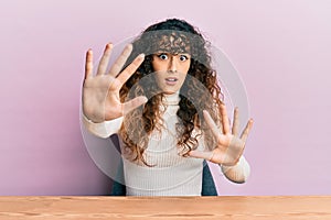 Young hispanic girl wearing casual clothes sitting on the table afraid and terrified with fear expression stop gesture with hands,