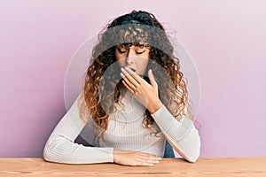 Young hispanic girl wearing casual clothes sitting on the table bored yawning tired covering mouth with hand