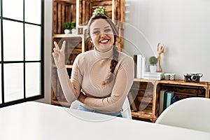 Young hispanic girl wearing casual clothes sitting on the table at home smiling with happy face winking at the camera doing