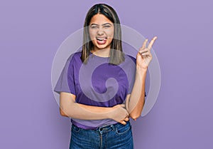 Young hispanic girl wearing casual purple t shirt smiling with happy face winking at the camera doing victory sign