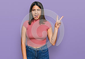 Young hispanic girl wearing casual t shirt smiling with happy face winking at the camera doing victory sign