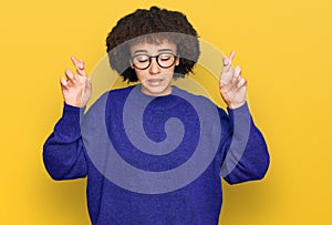 Young hispanic girl wearing casual winter sweater and glasses gesturing finger crossed smiling with hope and eyes closed