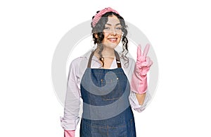 Young hispanic girl wearing cleaner apron and gloves smiling with happy face winking at the camera doing victory sign