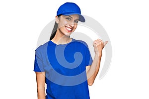 Young hispanic girl wearing delivery courier uniform smiling with happy face looking and pointing to the side with thumb up