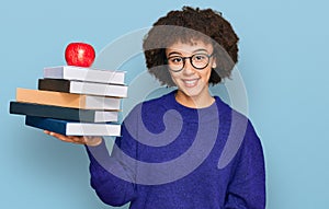 Young hispanic girl wearing glasses holding books and red apple looking positive and happy standing and smiling with a confident