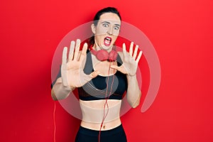 Young hispanic girl wearing gym clothes and using headphones afraid and terrified with fear expression stop gesture with hands,