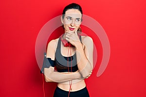 Young hispanic girl wearing gym clothes and using headphones with hand on chin thinking about question, pensive expression