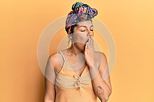 Young hispanic girl wearing hair turban over yellow background bored yawning tired covering mouth with hand