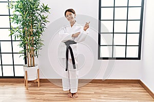 Young hispanic girl wearing karate kimono and black belt with a big smile on face, pointing with hand and finger to the side