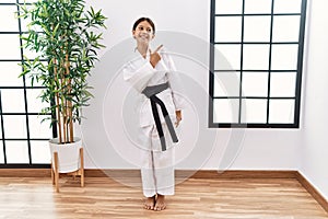 Young hispanic girl wearing karate kimono and black belt cheerful with a smile of face pointing with hand and finger up to the