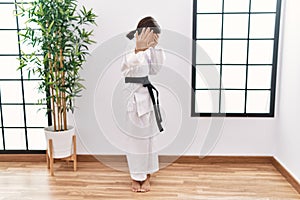 Young hispanic girl wearing karate kimono and black belt covering eyes with hands and doing stop gesture with sad and fear