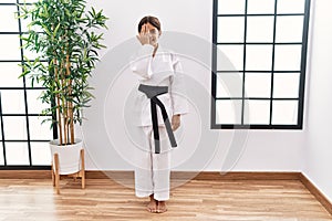 Young hispanic girl wearing karate kimono and black belt covering one eye with hand, confident smile on face and surprise emotion