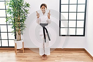 Young hispanic girl wearing karate kimono and black belt gesturing finger crossed smiling with hope and eyes closed