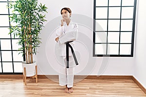Young hispanic girl wearing karate kimono and black belt with hand on chin thinking about question, pensive expression