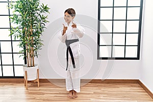 Young hispanic girl wearing karate kimono and black belt pointing fingers to camera with happy and funny face