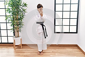 Young hispanic girl wearing karate kimono and black belt smelling something stinky and disgusting, intolerable smell, holding