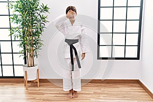 Young hispanic girl wearing karate kimono and black belt smiling and laughing with hand on face covering eyes for surprise