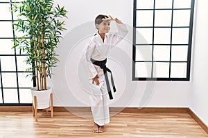 Young hispanic girl wearing karate kimono and black belt very happy and smiling looking far away with hand over head