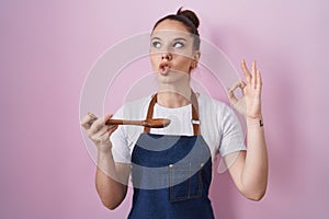 Young hispanic girl wearing professional cook apron holding wood spoon making fish face with mouth and squinting eyes, crazy and