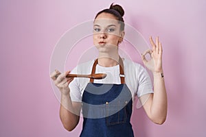 Young hispanic girl wearing professional cook apron holding wood spoon puffing cheeks with funny face