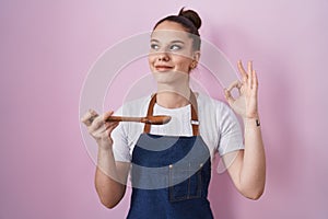 Young hispanic girl wearing professional cook apron holding wood spoon smiling looking to the side and staring away thinking