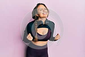 Young hispanic girl wearing sportswear and headphones excited for success with arms raised and eyes closed celebrating victory