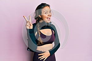 Young hispanic girl wearing sportswear and headphones smiling with happy face winking at the camera doing victory sign