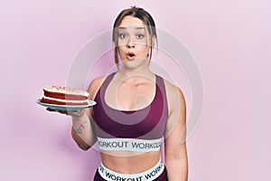 Young hispanic girl wearing sportswear holding carrot cake scared and amazed with open mouth for surprise, disbelief face