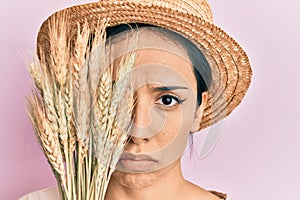 Young hispanic girl wearing summer hat holding spike wheat over eye depressed and worry for distress, crying angry and afraid