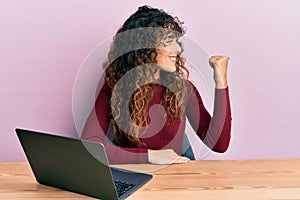 Young hispanic girl working using computer laptop pointing thumb up to the side smiling happy with open mouth