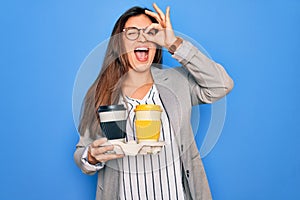 Young hispanic intern business woman holding coffee over blue background with happy face smiling doing ok sign with hand on eye