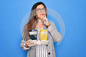 Young hispanic intern business woman holding coffee over blue background serious face thinking about question, very confused idea