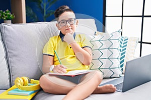 Young hispanic kid doing homework sitting on the sofa with hand on chin thinking about question, pensive expression