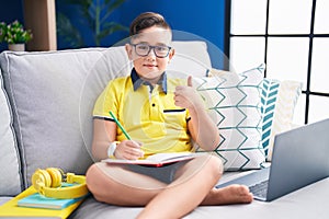 Young hispanic kid doing homework sitting on the sofa doing happy thumbs up gesture with hand