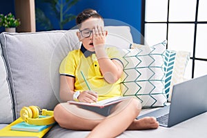 Young hispanic kid doing homework sitting on the sofa yawning tired covering half face, eye and mouth with hand