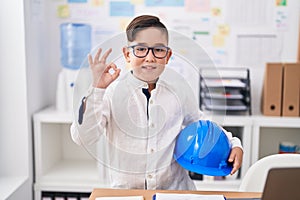 Young hispanic kid holding architect hardhat at the office doing ok sign with fingers, smiling friendly gesturing excellent symbol