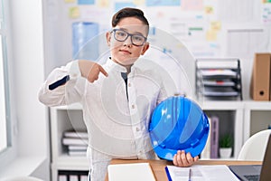 Young hispanic kid holding architect hardhat at the office pointing finger to one self smiling happy and proud