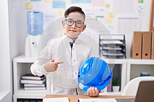 Young hispanic kid holding architect hardhat at the office smiling happy pointing with hand and finger