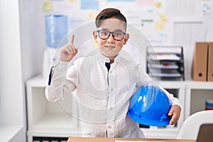 Young hispanic kid holding architect hardhat at the office surprised with an idea or question pointing finger with happy face,