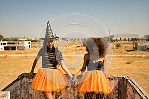 Young Hispanic and Latina women holding hands, smiling and leaning against a stone wall dressed for Halloween outside