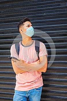 Young Hispanic male wearing a facemask and holding a phone while leaning on a garage wall