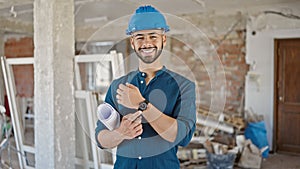 Young hispanic man architect wearing hardhat holding blueprints pointing at wrist watch at construction site