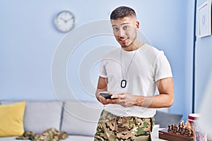 Young hispanic man army soldier using smartphone at home